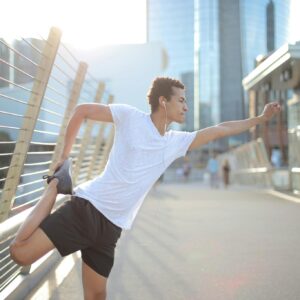 Young African American athlete in earbuds listening to music and stretching legs while training alone on street